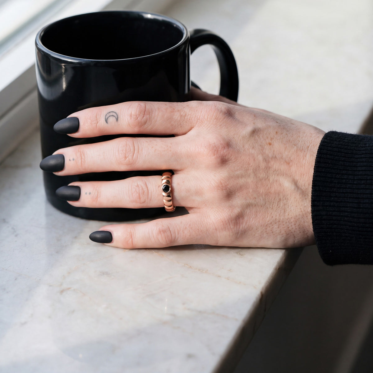 Rose gold scalloped wedding band with a black diamond on a hand beside a black coffee mug