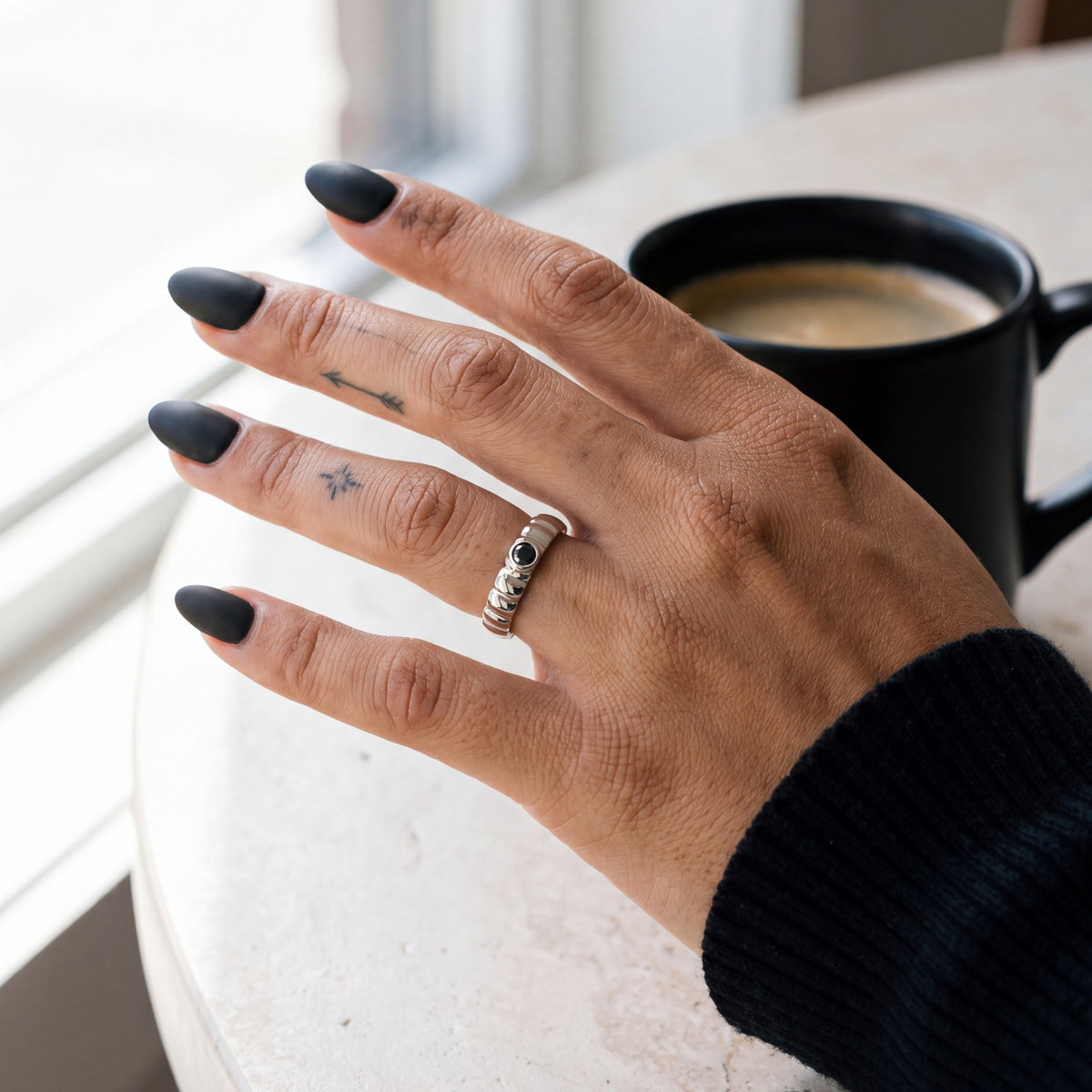 White gold Gravitational Wave wedding band with a black diamond on a hand beside a black coffee mug