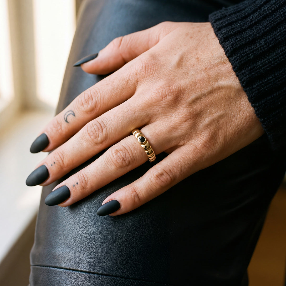 Yellow gold scalloped wedding band with a black diamond on a hand resting on black leather trousers