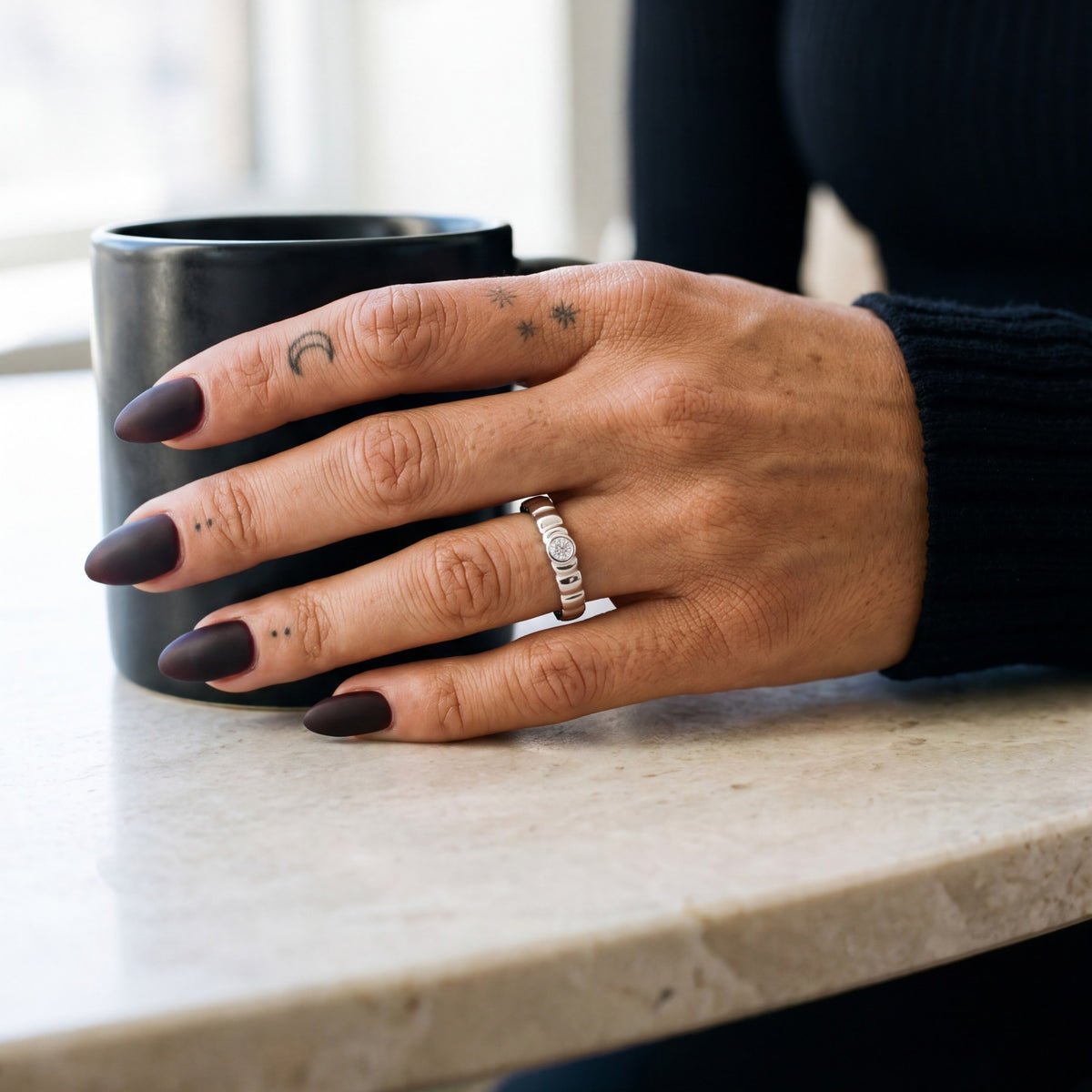 White gold Gravitational Wave band with a white diamond on a hand beside a black coffee mug